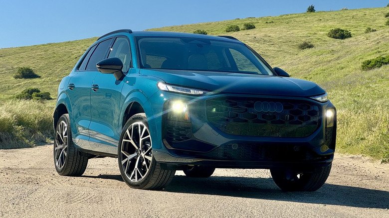 A front three quarters shot of the blue Audi Q3 with its headlights on parked on a dirt road in front of a grass and shrub-covered hill A front three quarters shot of the blue Audi Q3 with its headlights on parked on a dirt road in front of a grass and shrub-covered hill